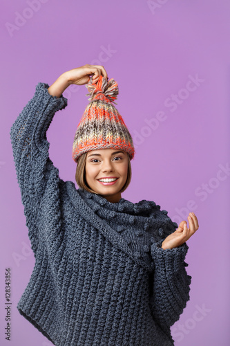 Young funny beautiful fair-haired girl in knited hat and sweater smiling looking at camera posing over violet background. Copy space.