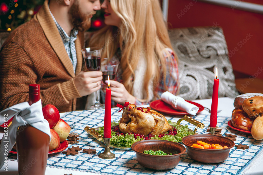 Beautiful couple kissing and holding glass of wine in a decorated festive interior with a Christmas tree. A romantic dinner for thanksgiving with fried chicken and candles