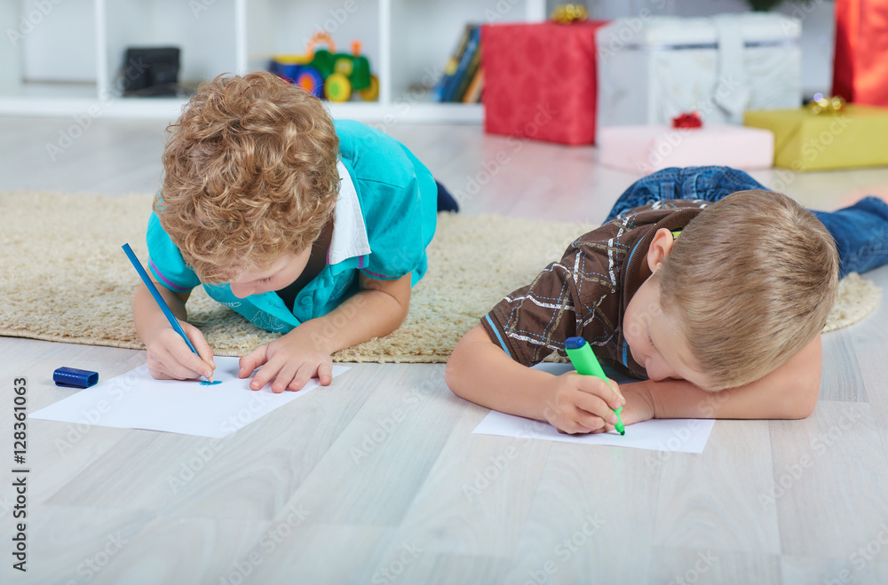 Two boys draw Santa Claus on the paper on the floor in the nursery. Boxes with gifts in the background.