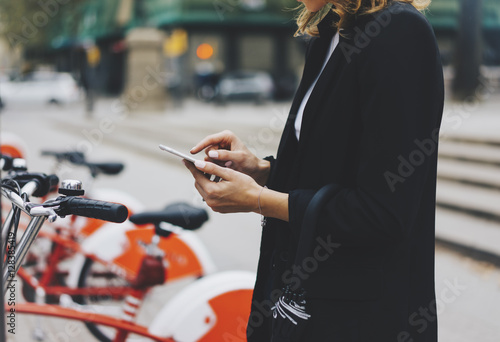 Young businesswomen in black suit and umbrella using smartphone, biking and going to work by city bicycle on urban street, hipster girl holding mobile gadget, ecology environment concept