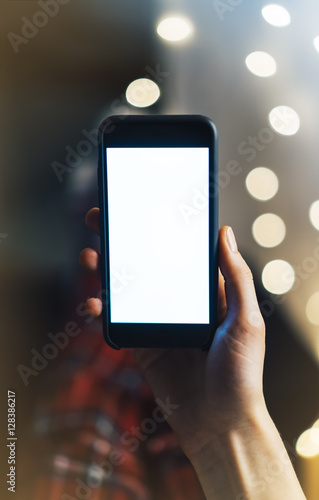 Girl pointing finger on smartphone with empty blank screen monitor on the background bokeh light in home atmosphere, hipster using in female hands smart phone with blank template, christmas blur