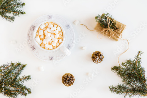 Winter Christmas decoration, fir branches and cones, wrapped gift, marshmallows and hot drink, coffee or cacao in a cup. White table, top view, flat lay.