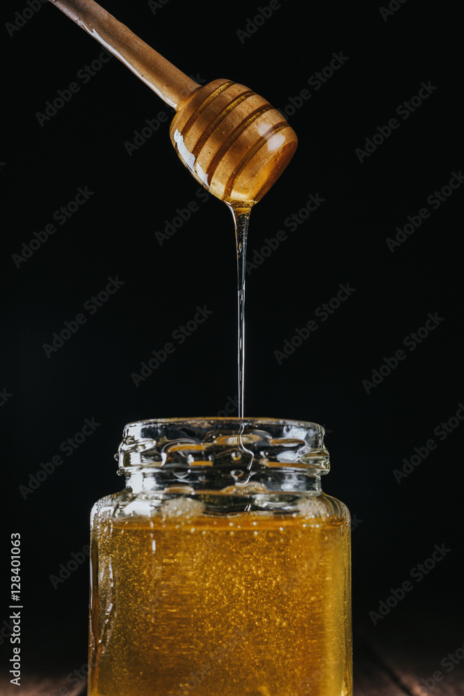 Crop hand dipping wooden honey stick in glass jar of honey isolated on black . Crop hand dipping wooden honey stick in glass jar of honey isolated on black .