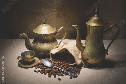 Heap of coffee beans in burlap sack with a vintage spoon and different vintage turkish coffeepots on canvas background photo