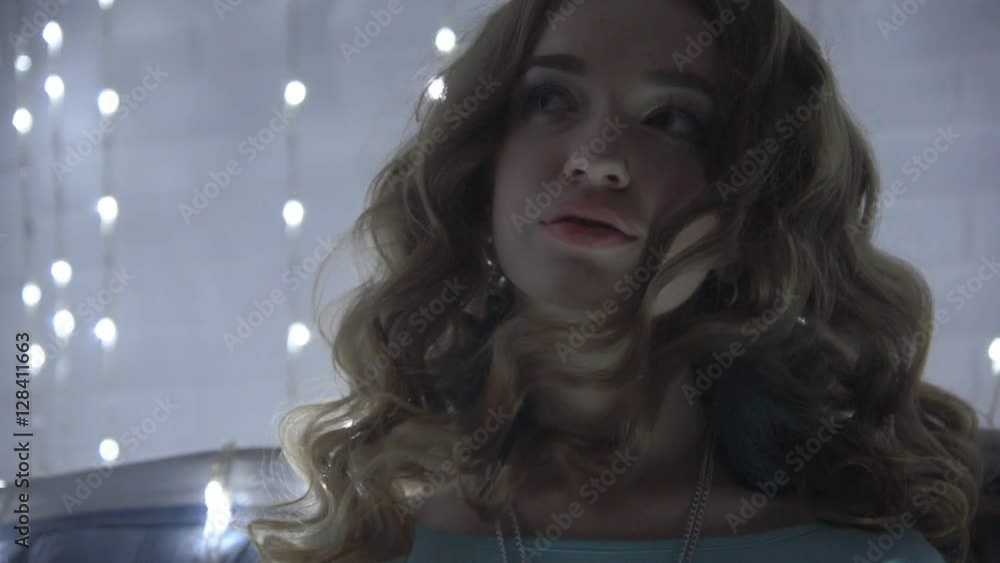 Brunette woman in a turquoise dress sitting on a luxury sofa near white brick wall decorated with garland. A festive portrait of a girl with a christmas light bulbs. Female hair develop from the wind. Brunette woman in a turquoise dress sitting on a luxury sofa near white brick wall decorated with garland. A festive portrait of a girl with a christmas light bulbs. Female hair develop from the wind.