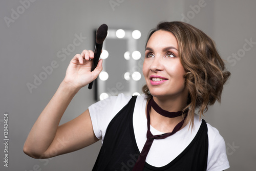 Portrait of a beautiful woman holding a makeup brush powder and looking up. Mirror with lights on background