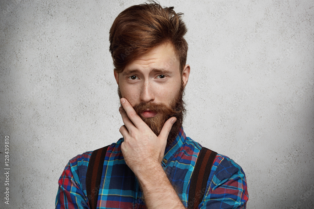 Studio portrait of fashionable bearded man wearing flannel shirt and  suspenders having doubtful and quizzical look, touching his thick beard as  thinking about something while posing against blank wall Stock Photo