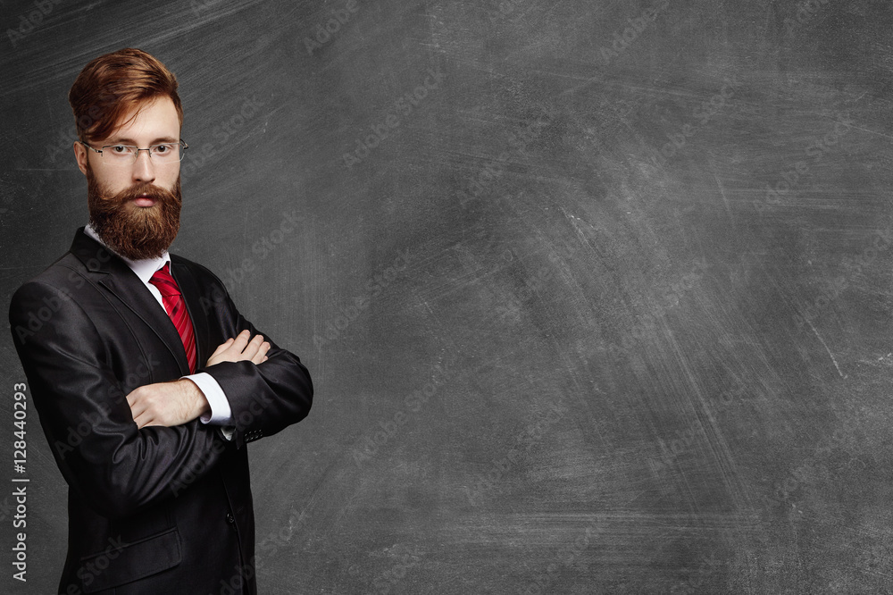 Stylish young bearded professor in glasses standing at blank chalkboard, keeping his arms crossed and looking at camera with serious or angry expression. Businessman in formal wear posing indoors Stylish young bearded professor in glasses standing at blank chalkboard, keeping his arms crossed and looking at camera with serious or angry expression. Businessman in formal wear posing indoors