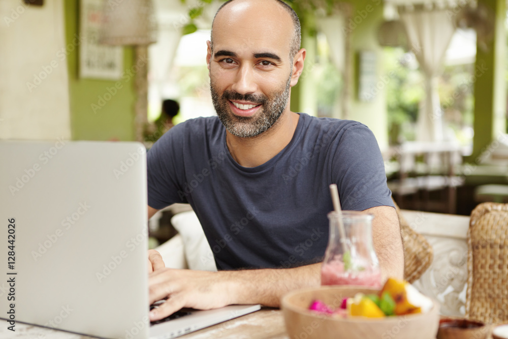 People, lifestyle and modern technology. Outdoor portrait of happy handsome man sitting at cafe table with glass of smoothie and food during breakfast, keeping hands on keyboard of his generic laptop People, lifestyle and modern technology. Outdoor portrait of happy handsome man sitting at cafe table with glass of smoothie and food during breakfast, keeping hands on keyboard of his generic laptop