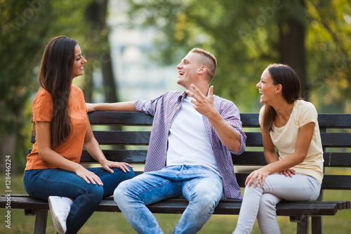 Three friends are sitting on bench in park and talking. 