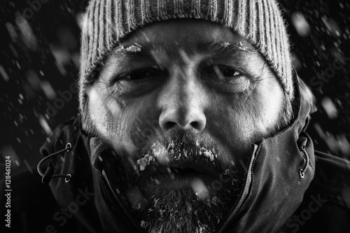 Freezing cold man standing in a snow storm blizzard trying to keep warm. Wearing a beanie hat and winter coat with frost and ice on his beard and eyebrows staring at the camera. Black and white. photo