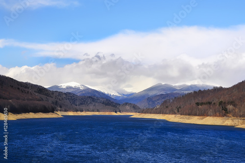 blue lake at the foot of the mountains covered with snow  beautiful clouds and sky  the forest slopes. Beautiful winter landscape  the beginning of spring.