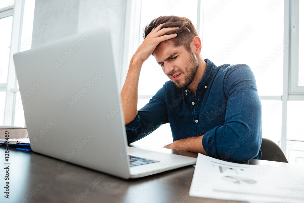 Confused young man sitting near laptop and holding head Confused young man sitting near laptop and holding head