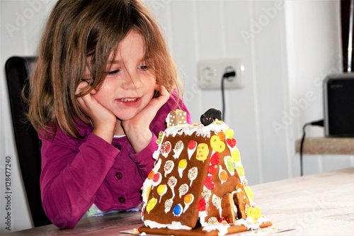Ein kleines Mädchen sitzt am Küchentisch in der Küche vor einem selbstgemachten Lebkuchenhaus und freut sich.