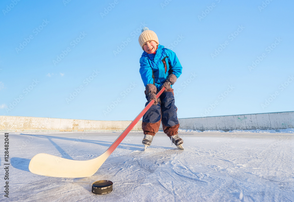 Fototapeta premium Winter games. Young happy child playing ice hockey on an outdoor ice rink. Focus on the boy.