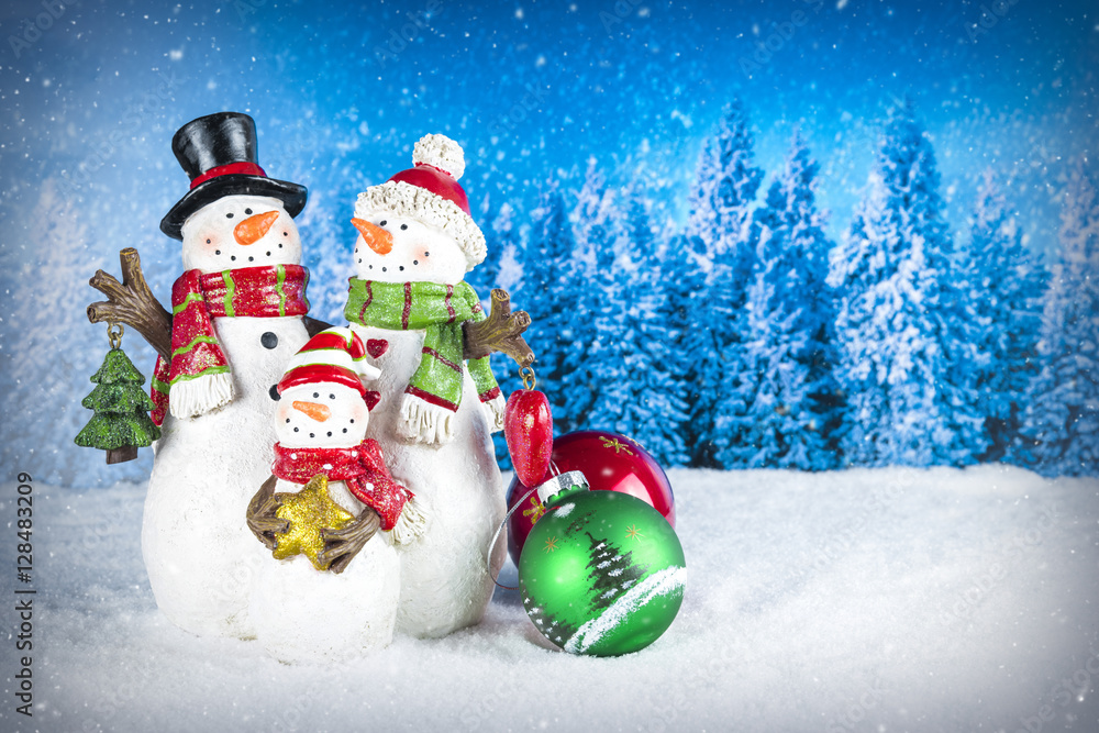 Fototapeta premium Christmas decoration with snowman figurines as a family, colorful baubles on a blue snow pine tree landscape background.
