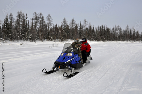 Two men ride on a snowmobile on the frost river in the winter forest
