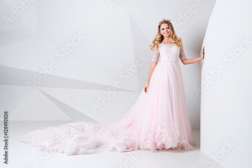Beautiful blonde woman wearing gorgeous long dress and crystal crown posing in studio on white polygonal background.