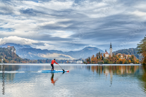 Stand up male paddle surfer training at Bled lake in Slovenia. Beautiful Alps mountain and castle on island at background. Autumn season, forest with fall yellow leaves trees at background. 