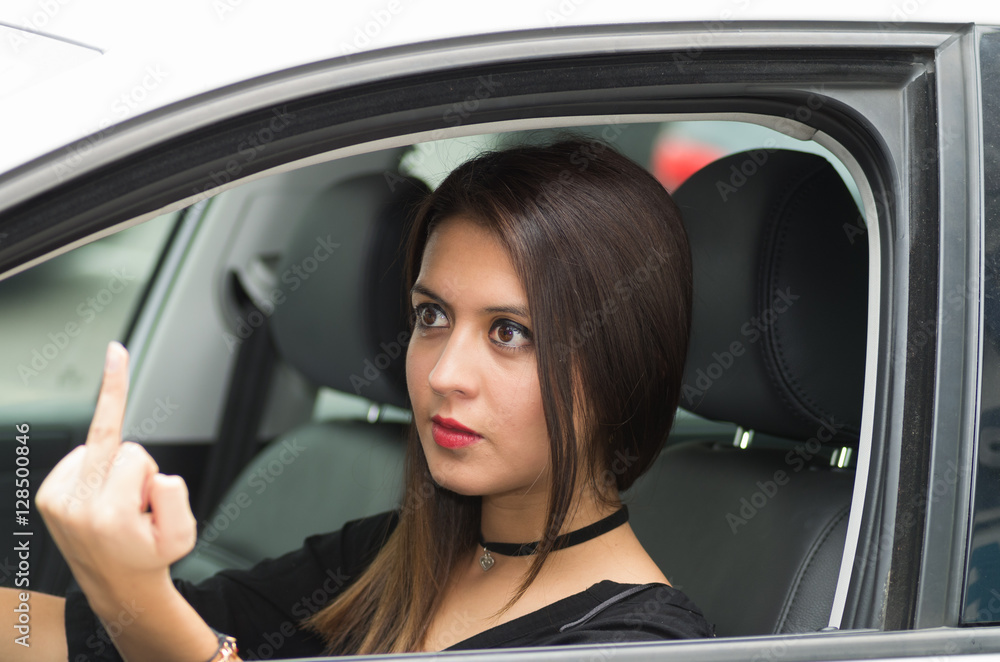 Closeup young woman sitting in car giving the finger angrily, as seen from outside drivers window, female driver concept