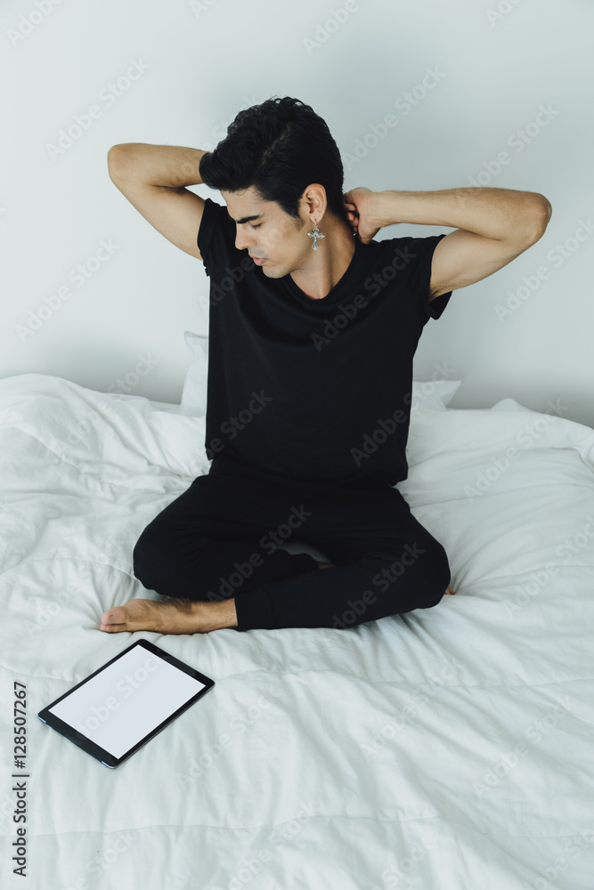Young handsome attractive brazilian male in black t-shirt and tights stretching his arms sitting on bed after sleeping in the morning, mock up of digital tablet with white screen laying near him Young handsome attractive brazilian male in black t-shirt and tights stretching his arms sitting on bed after sleeping in the morning, mock up of digital tablet with white screen laying near him