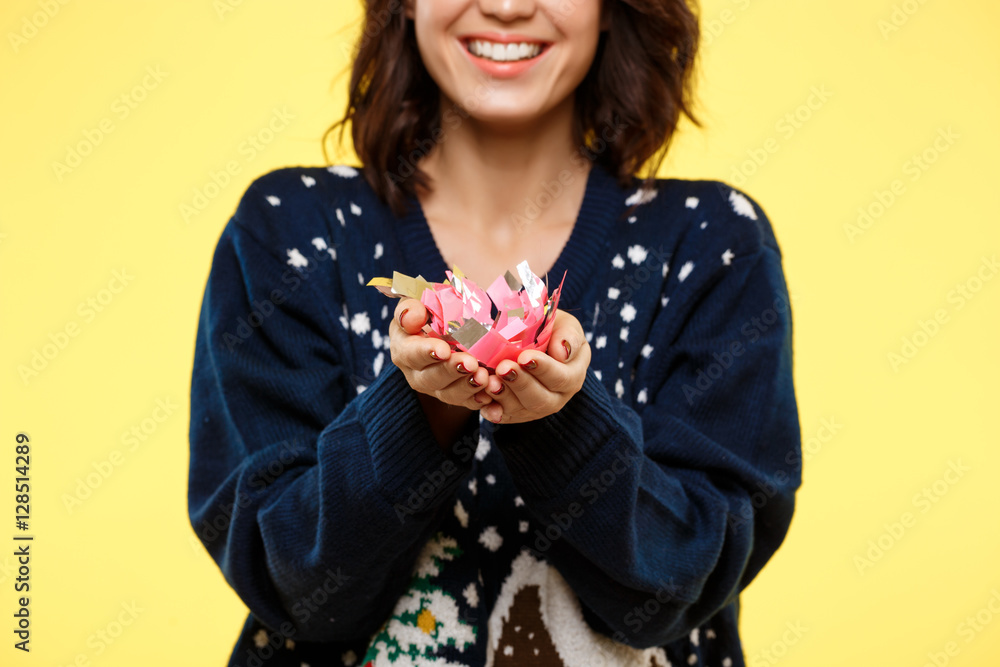Young cheerful beautiful brunette girl in cosy knited sweater smiling with handful of confetti over yellow background. Young cheerful beautiful brunette girl in cosy knited sweater smiling with handful of confetti over yellow background.