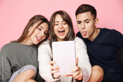 Young beautiful friends making selfie on tablet over pink background.