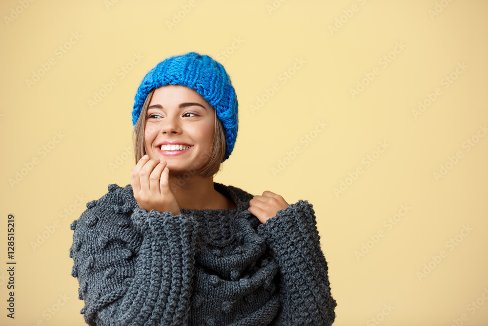 Young beautiful fair-haired girl in knited hat and sweater smiling looking at side over yellow background. Young beautiful fair-haired girl in knited hat and sweater smiling looking at side over yellow background.