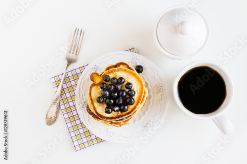 Stack of homemade pancakes with black currant, vintage plate and fork, cup of coffee, white background, top view.