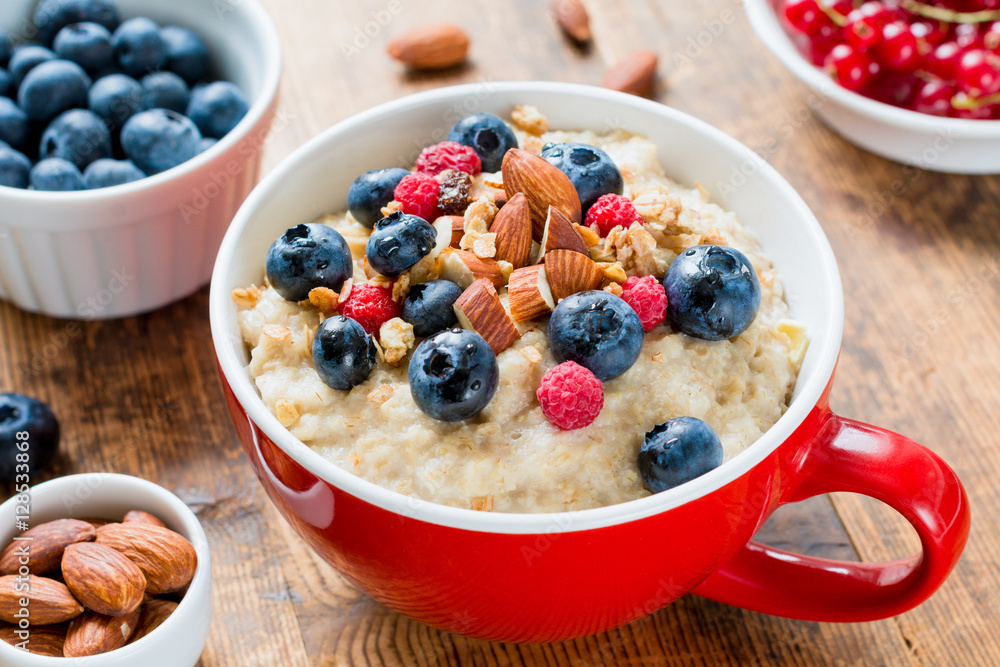 Close up view of delicious breakfast oatmeal porridge in red bowl topped with fresh blueberries, raspberries, almonds and granola. Close up view of delicious breakfast oatmeal porridge in red bowl topped with fresh blueberries, raspberries, almonds and granola.