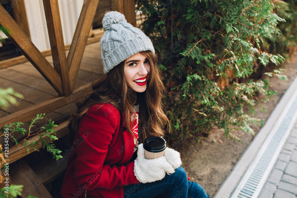 Portrait pretty girl in red coat, knitted hat sitting on wooden stairs near green branches outdoor. She holds coffee in white gloves and smiling to camera. View from above Portrait pretty girl in red coat, knitted hat sitting on wooden stairs near green branches outdoor. She holds coffee in white gloves and smiling to camera. View from above