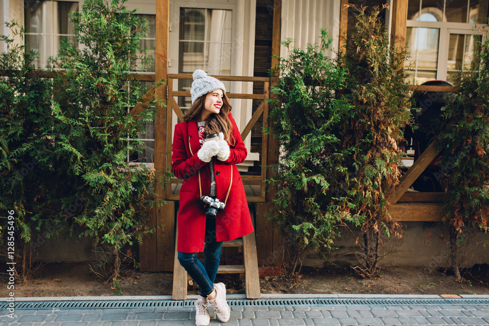 Full length pretty girl with long hair in red coat and knitted hat standing on wooden house background. She holds camera and coffee to go in white gloves, smiling to side