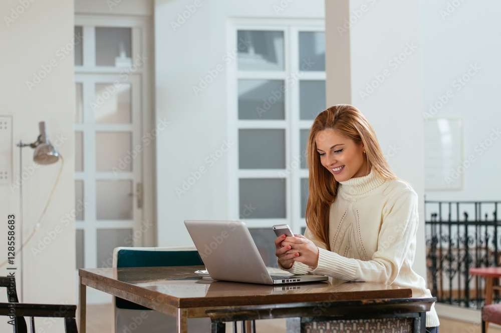 Attractive young woman looking at the mobile phone and smiling while sitting at her working place in office Attractive young woman looking at the mobile phone and smiling while sitting at her working place in office
