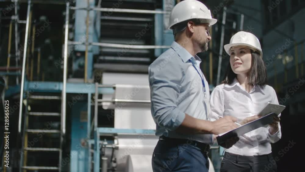 Female executive engineer in hard hat holding tablet and showing bearded male colleague with clipboard factory workshop area Female executive engineer in hard hat holding tablet and showing bearded male colleague with clipboard factory workshop area