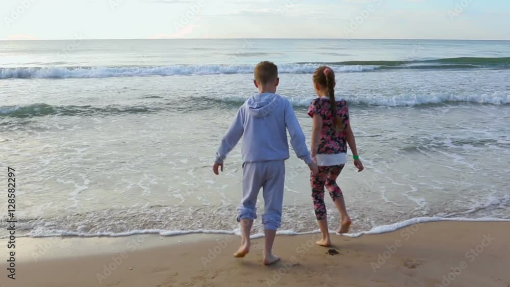 Children Hold Hands And Run Toward Ocean, Stop, And Feel The Waves Crash On Their Feet Children Hold Hands And Run Toward Ocean, Stop, And Feel The Waves Crash On Their Feet