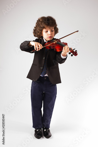 Smart little boy in a business suit concentrating playing the violin. Gray background.