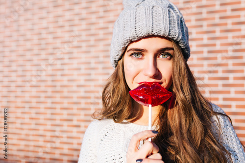 Closeup portrait young girl with long hair in knitted hat with caramel red lips on wall background outside. She is looking to camera
