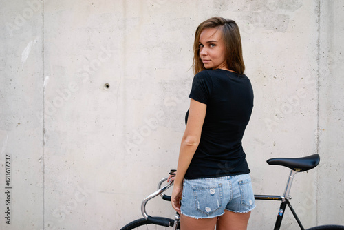 A brunette girl with long hair wearing a blank black t-shirt is standing with the back to the camera on a gray wall background on a street. Empty space for text or design.