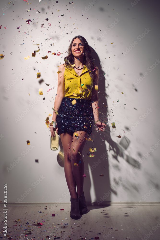 Drunk happy girl holding a bottle of Champagne and standing close to the wall. Confetti in the air. Home party Drunk happy girl holding a bottle of Champagne and standing close to the wall. Confetti in the air. Home party