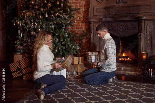 Merry Christmas. Young couple celebrating New Year at home. Man and woman in knitted sweater sitting on the floor by fireplace