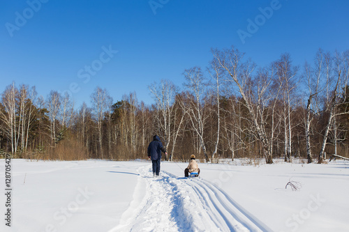 Dad carries daughter sledging in winter frosty day in the woods. After sled trace remains.