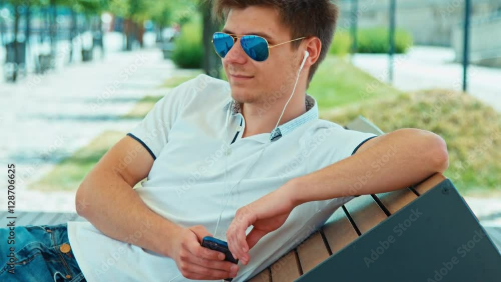 Beautiful student male in sunglasses using mobile phone with headphones sitting on outdoor bench and smiling Beautiful student male in sunglasses using mobile phone with headphones sitting on outdoor bench and smiling