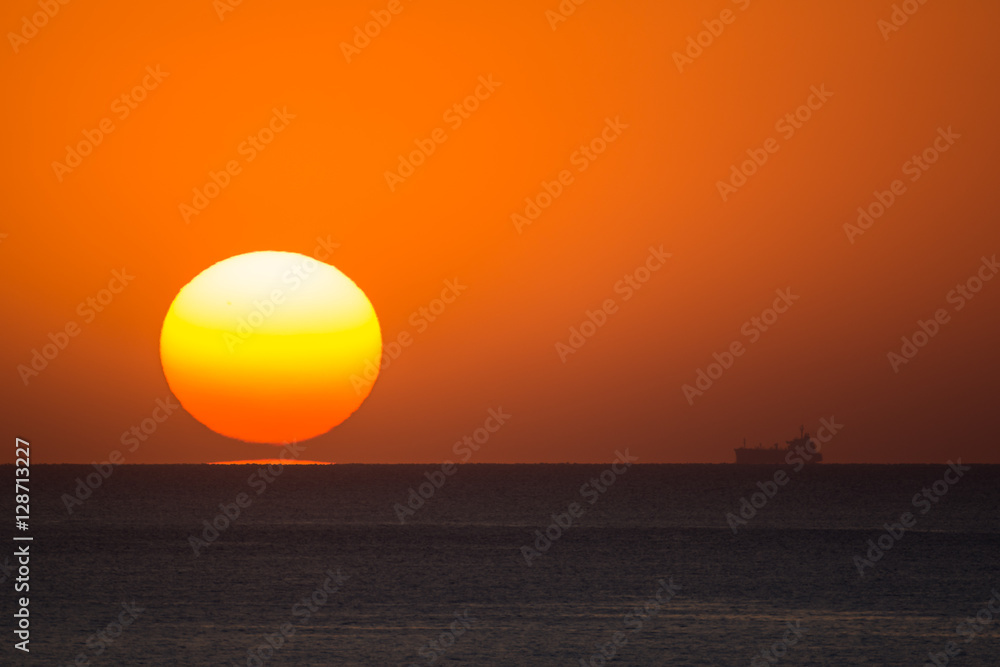 Sun sets near a fade silhouette of a ship on the horizon