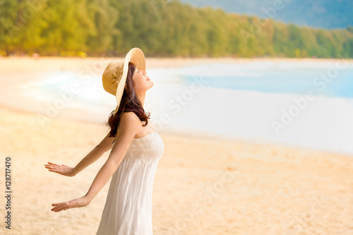 Healthy woman open arms breathing deep at sunrise beach.
Woman in white dress with sunhat inhaling fresh air and sunbathe a morning light at beautiful  beach  

