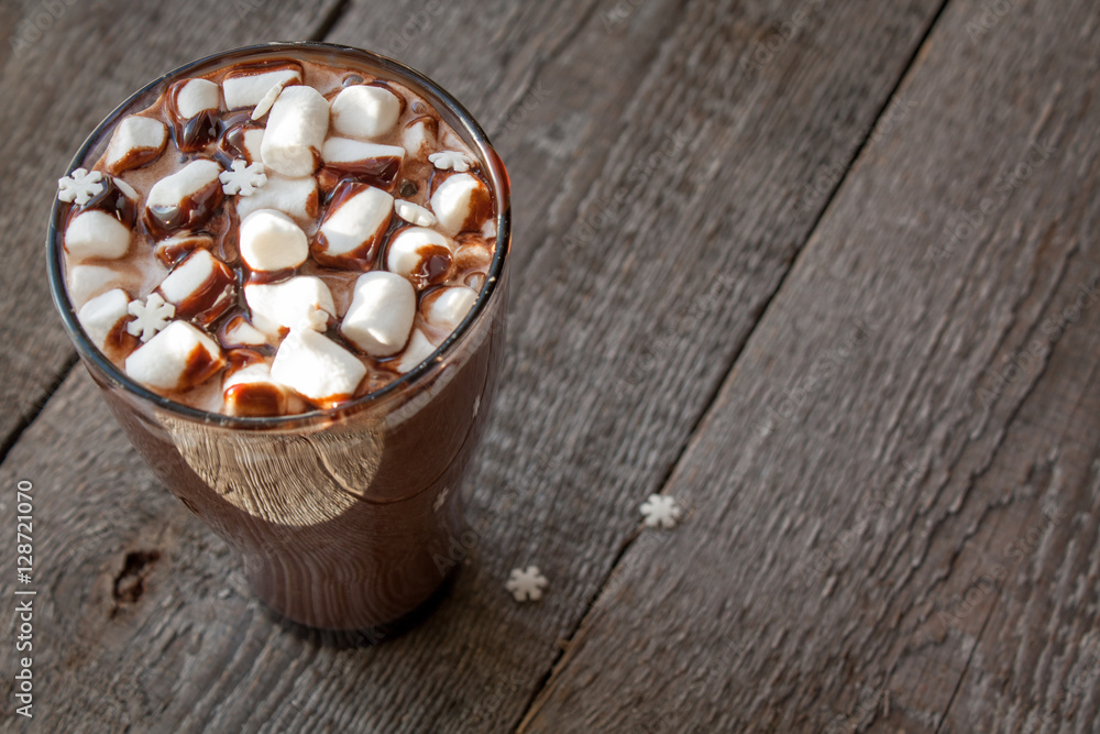 Winter hot cocoa with marshmallows, chocolate syrup, sugar snowflakes in a tall glass on a wooden background. Winter hot cocoa with marshmallows, chocolate syrup, sugar snowflakes in a tall glass on a wooden background.