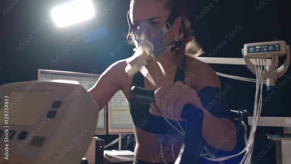 Tilt up of strong woman in oxygen mask cycling exercise bike in darkly lit laboratory