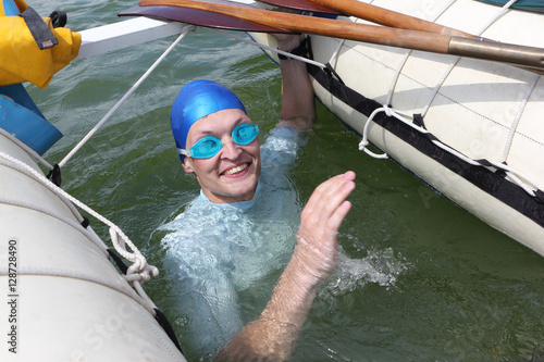 The young man swimming in the river in a blue cap and goggles     near a catamaran  
