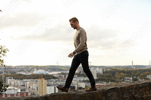 Man wearing a brown sweater and jeans, walking along a mountain top with the great cityview behind him on a summer day. Looking away from camera.