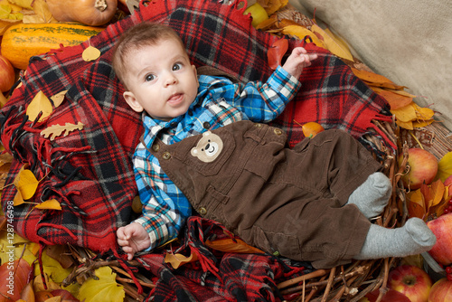 autumn little boy lie on plaid blanket, yellow fall leaves, apples, pumpkin and decoration on textile