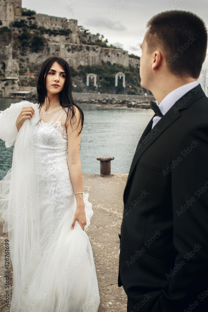 Bride holds her dress and looks at groom while they stand on the